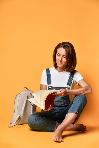 Portrait of a pensive girl in a white t-shirt and denim overalls, sitting on the floor with a crown pad and a pencil on a yellow background - Foto, immagini
