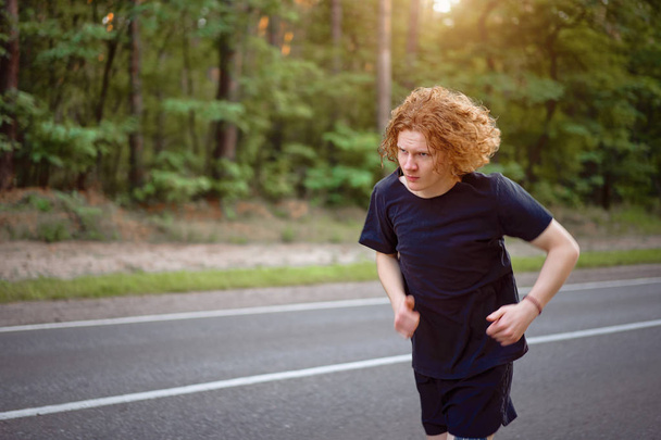 Caucasian young curly man athlete runs sunny summer day on asphalt road in the forest. - Valokuva, kuva