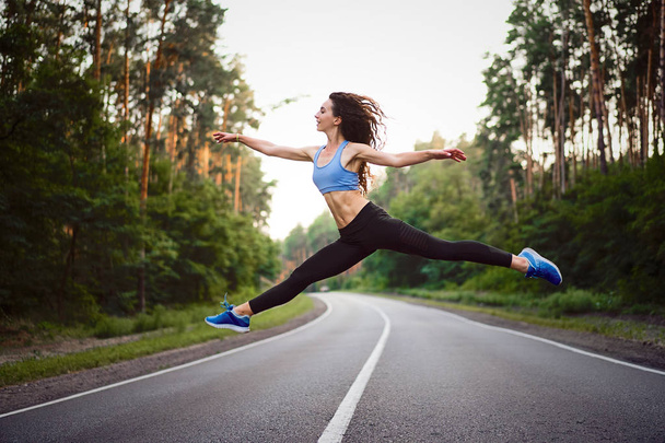 young beautiful brunette girl jumping in twine with background  asphalt road and pine forest - Фото, изображение