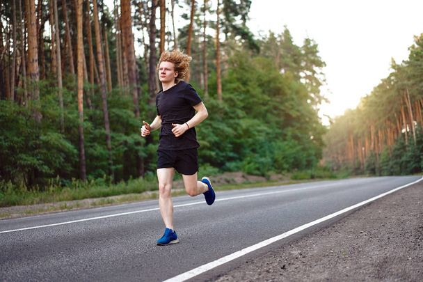 Valkoihoinen nuori kihara mies urheilija kulkee aurinkoinen kesäpäivä asfalttitiellä metsässä
. - Valokuva, kuva