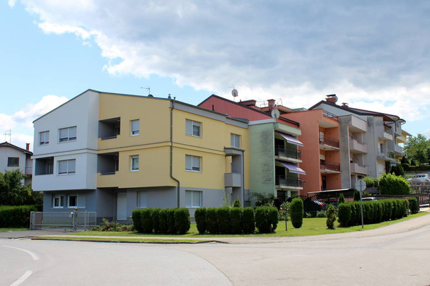 Bunte Einfamilienhäuser am Stadtrand, umgeben von Metallzäunen und ungeschnittenen Hecken neben asphaltierter Straße am wolkenverhangenen, blauen Himmel im Hintergrund - Foto, Bild