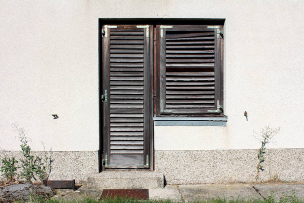 Omakotitalon sisäänkäynti ovet suljettu rappeutunut puinen ikkuna kaihtimet haalistunut väri asennettu vaaleanharmaa julkisivu ympäröi betoni polku ja leikkaamaton ruoho lämmin aurinkoinen kesäpäivä
 - Valokuva, kuva