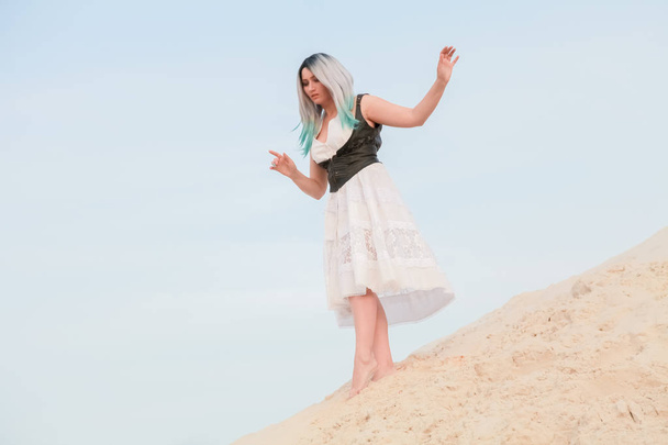 Young beautiful Caucasian woman in white dress and brown leather waistcoat posing in desert landscape with sand. - Photo, Image