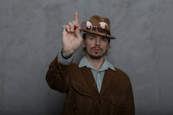 Old-fashioned strong young man in a vintage hat in a round glasses in a stylish brown jacket stands near a gray wall in the studio. Brutal hipster guy draws attention raises his index finger up. - Fotoğraf, Görsel