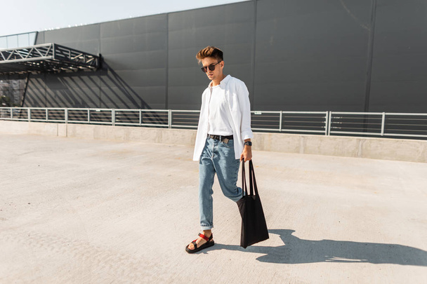 Modern young man hipster in trendy white and denim clothes in sunglasses in red sandals with a vintage bag walks on the street on a sunny day. Urban guy on vacation. Summer stylish men's clothing. - Photo, Image