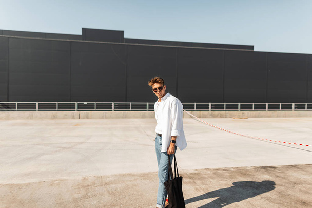 Ein urbaner junger Hipster mit stylischer Frisur in modischer Sommerkleidung und schwarzer Stofftasche mit Vintage-Sonnenbrille spaziert an einem sonnigen Tag in der Nähe eines grauen Gebäudes durch die Stadt. Europäischer Typ.  - Foto, Bild