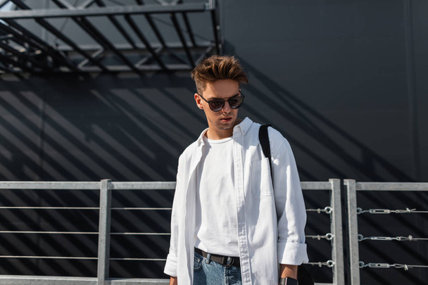 Modieuze jonge man model in een T-shirt in een stijlvolle witte shirt in blauwe jeans in zonnebril wandelingen rond de stad in de buurt van een grijs gebouw. Europese hipster man geniet zonnige dag. Streetstyle. - Foto, afbeelding