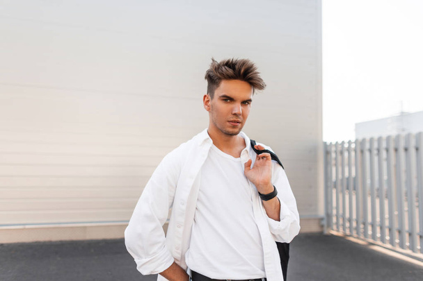 Şık siyah bez çanta ile şık siyah bez çanta ile şık beyaz giysiler yakışıklı modern genç adam şehirde beyaz bir binanın yakınında duruyor. Amerikalı adam model açık havada. Yaz stili. - Fotoğraf, Görsel