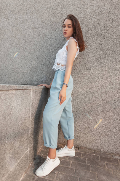 Pretty fashionable young woman in stylish summer clothes in white leather trendy sneakers stands near a gray building in the city. Attractive european brunette girl outdoors. Youth style. - Foto, immagini