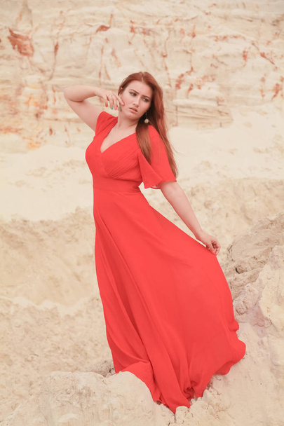 Young beautiful Caucasian woman in long red dress posing in desert landscape with sand. - Photo, Image
