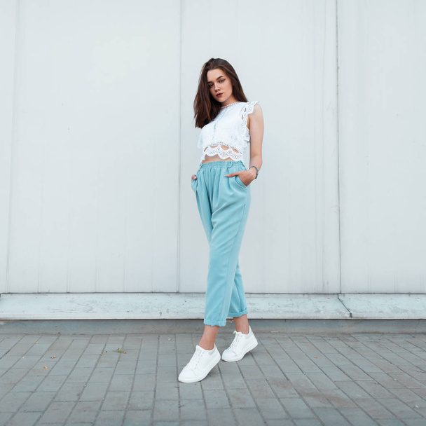 Pretty stylish young woman in fashionable summer clothes in leather white sneakers posing near a vintage wall on a street in the city. Attractive girl model outdoors. Trendy clothes. New collection. - Photo, Image