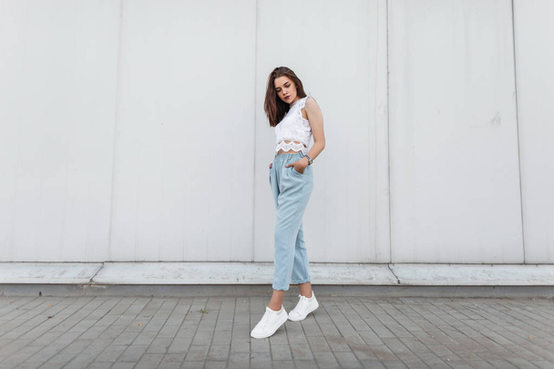 Modern beautiful young woman in a fashionable lacy t-shirt in vintage blue pants in white sneakers stands near a wall in the city. Pretty attractive brunette girl. Elegant women's youth clothing. - Foto, Bild
