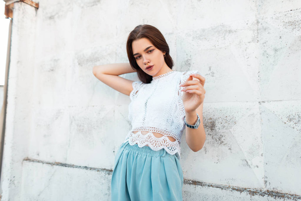 Cute attractive young woman straightens hair. Stylish beautiful girl in trendy lace top in fashionable blue pants poses on the street near the vintage wall. New collection of summer women's clothing. - Foto, afbeelding