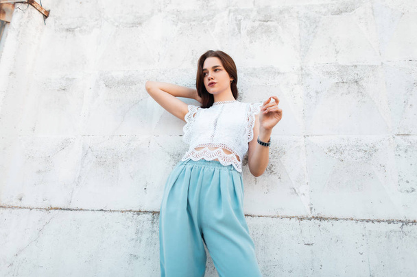 Fashionable beautiful young woman in elegant clothes posing near the vintage white wall on the street. European pretty stylish girl fashion model. Trendy summer women's clothing. - Fotó, kép