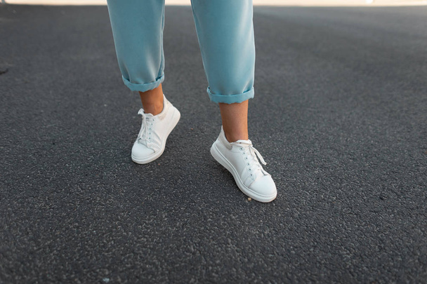 Female legs in stylish blue pants in white leather sneakers on the asphalt in the city. Trendy young woman on a walk. Fashionable women's shoes. New collection.  - Foto, imagen