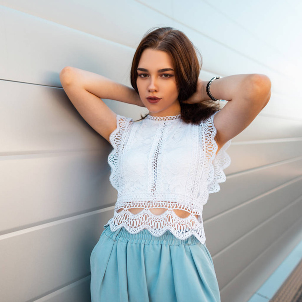 Pretty attractive young woman straightens hair. European urban girl in trendy lace top in fashionable blue pants poses on the street near the vintage wall. New collection of summer women's clothing. - Fotografie, Obrázek
