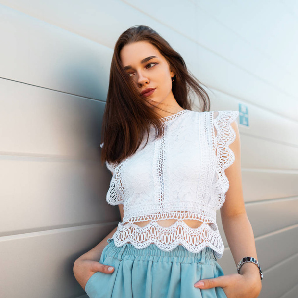Jonge elegante vrouw fashion model in een modieuze Lace top in stijlvolle blauwe broek staat in de stad in de buurt van een witte Vintage muur. Moderne trendy meisje rusten in de straat. Zomer stijl. - Foto, afbeelding