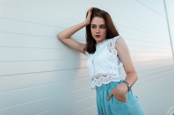 Melko kaunis nuori brunette nainen tyylikäs valkoinen pusero vintage sininen housut lepää lähellä vintage seinä kaupungissa. Moderni kaupunkilaistyttö rentoutuu ulkona kadulla. Kesätyyli
. - Valokuva, kuva