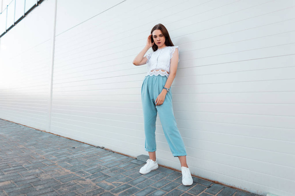 白いスニーカーの青いパンツのスタイリッシュなレーストップの美しい若い女性モデルは、市内の近代的な建物の近くに立っています。屋外でかわいい女の子。婦人服のトレンディな夏のコレクション. - 写真・画像