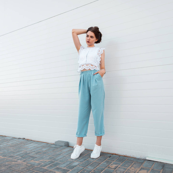 Pretty fashionable young woman model in stylish summer clothes in white leather fashionable sneakers stands and straightens hair near a white building outdoors. European brunette girl. Youth style. - Foto, Imagem