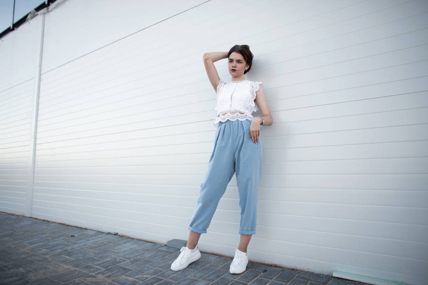 Modern beautiful young woman in a fashionable lacy t-shirt in trendy blue pants in leather white sneakers stands near a wall in the city. Pretty attractive brunette girl. Elegant women's clothing. - Φωτογραφία, εικόνα