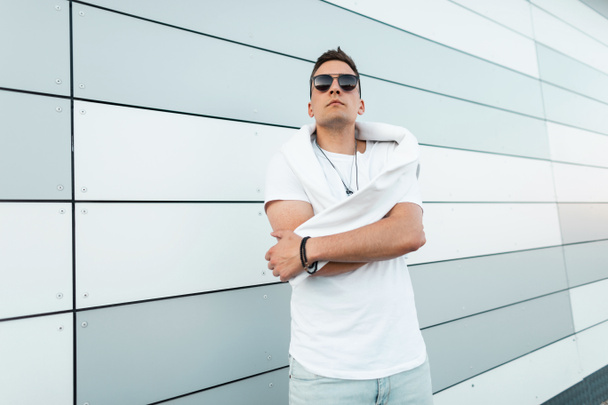 Fashionable young man fashion model with white sweatshirt in a stylish T-shirt in trendy black sunglasses posing near a modern wall in the city. American handsome hipster guy resting outdoors. - Photo, image
