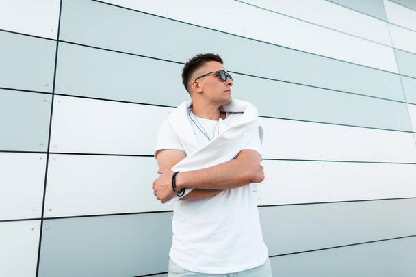 Κομψό ελκυστικό νεαρό χίππις σε ένα λευκό μοντέρνο μπλουζάκι με σκούρα γυαλιά ηλίου με ένα λευκό πουλόβερ που ποζάρει σε εξωτερικούς χώρους στην πόλη κοντά σε ένα σύγχρονο τοίχο. Αμερικανός τύπος. Καλοκαίρι νέα συλλογή άνδρες. - Φωτογραφία, εικόνα