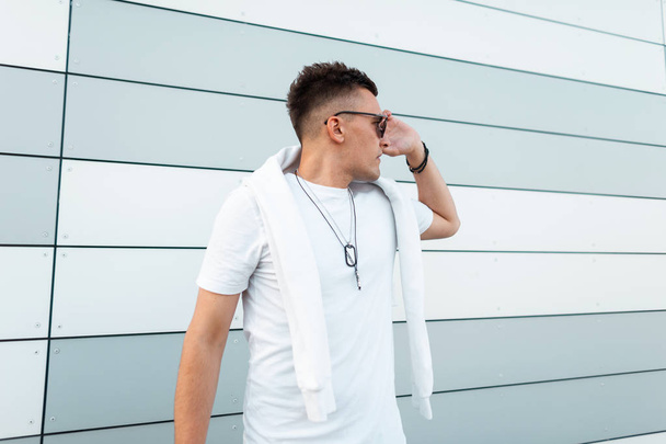 Urbano bonito jovem hipster homem em uma camiseta branca na moda em óculos de sol elegantes com penteado elegante posando perto de um edifício moderno na cidade. Modelo de homem atraente ao ar livre. Estilo de verão
. - Foto, Imagem