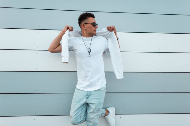 Σύγχρονος Αμερικανός νεαρός χίπστερ που φοράει γυαλιά ηλίου σε ένα μοντέρνο λευκό μπλουζάκι με μπλε τζιν ντύνεται ένα πουλόβερ κοντά σε ένα σύγχρονο ριγέ κτήριο στην πόλη μια καλοκαιρινή μέρα. Κουλ τύπος μόδας μοντέλο.  - Φωτογραφία, εικόνα