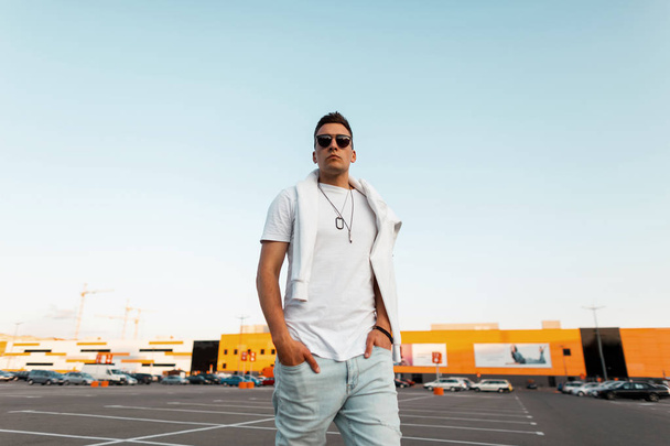 Stylish young man hipster with hairstyle in sunglasses in a fashionable white t-shirt with a sweatshirt in vintage blue jeans stand in the city on a summer day. American guy fashion model outdoors. - Fotografie, Obrázek