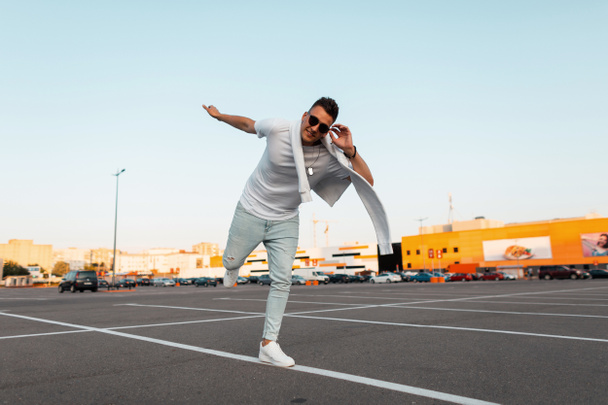 Positive young hipster man with smile in stylish white and jeans clothes in sunglasses in trendy sneakers stands on one foot in the city parking lot. Joyful guy keeps balance. Youth summer style. - Photo, Image
