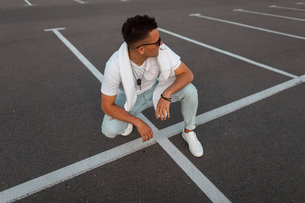 Young modern trendy hipster man in stylish dark sunglasses in vintage blue jeans in a fashionable white t-shirt in sneakers relaxes on a asphalt. Handsome guy model rests outdoors. Summer street style - Photo, Image