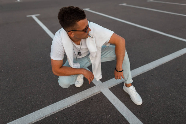 Amerykański model młodego mężczyzny w modne okulary przeciwsłoneczne w stylu Vintage niebieskie dżinsy w stylowym białym t-shirt w tenisówki siedzi na chodniku. Urban facet Hipster odpoczynku na zewnątrz. Letnia moda młodzieżowa. - Zdjęcie, obraz