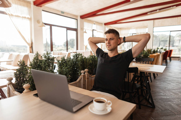 Succès joyeux jeune homme d'affaires dans un t-shirt noir avec un ordinateur se trouve dans un café moderne. Heureux pigiste travaillant à distance sur un ordinateur portable. Détendez-vous
. - Photo, image