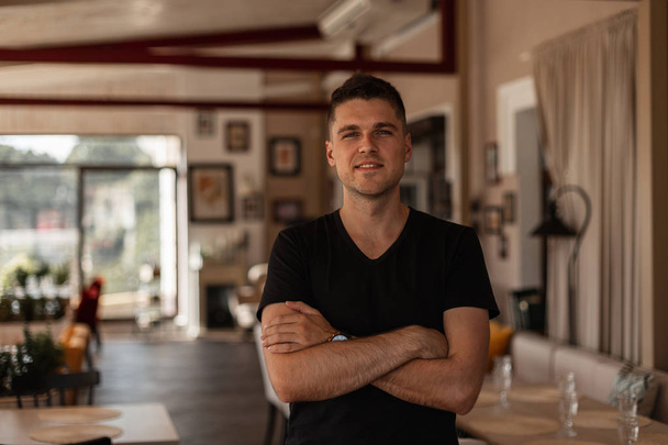 Urban european young man with trendy hairstyle in a stylish black t-shirt stands and smiles in a vintage cafe in a summer day. Handsome happy guy is resting indoors. Fashionable menswear. - 写真・画像