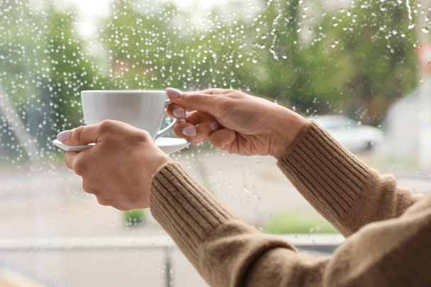 Young woman with cup of coffee near window indoors on rainy day, closeup - Foto, Imagen