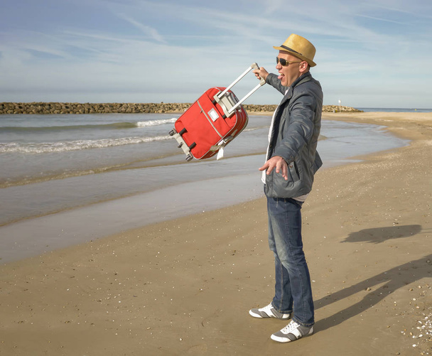 Felice giovane turista maschio gioisce in piedi in riva al mare, solleva una valigia rossa
 - Foto, immagini