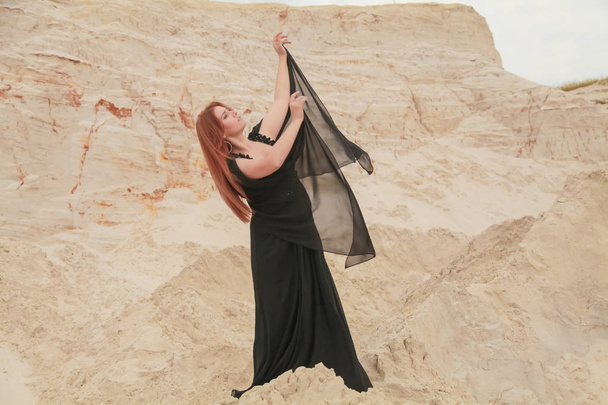 Jonge mooie blanke vrouw in zwart chiffon jurk poseren in woestijn landschap met zand. - Foto, afbeelding