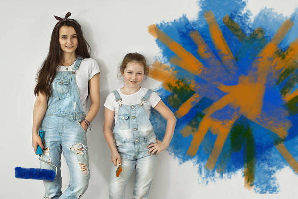 The girls paint the walls. Friendly collaboration between the two sisters. Teens in blue overalls make repairs together. The wall is partially painted with blue and orange paint. - Valokuva, kuva