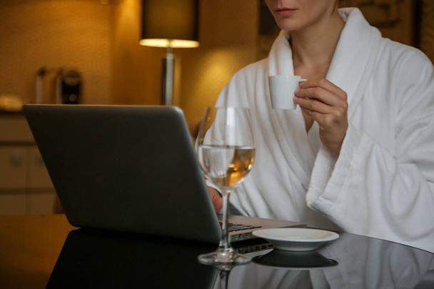 Frau im weißen Bademantel arbeitet am Laptop, entspannt nach einem langen Tag und verbringt einen gemütlichen Abend allein  - Foto, Bild