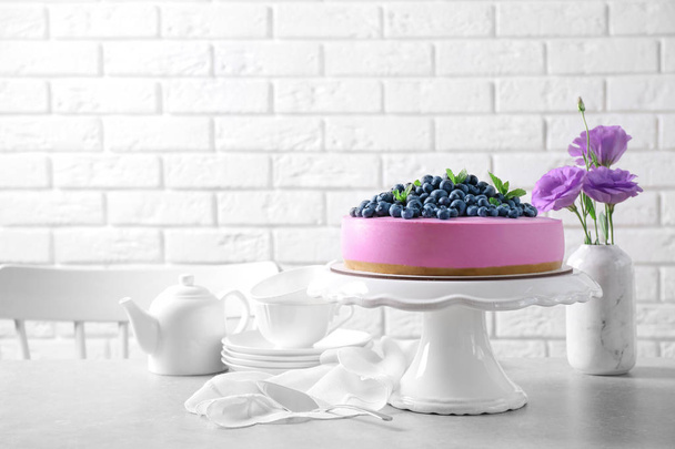 Soporte con sabroso pastel de arándanos y flores en la mesa gris contra la pared de ladrillo blanco. Espacio para texto
 - Foto, Imagen