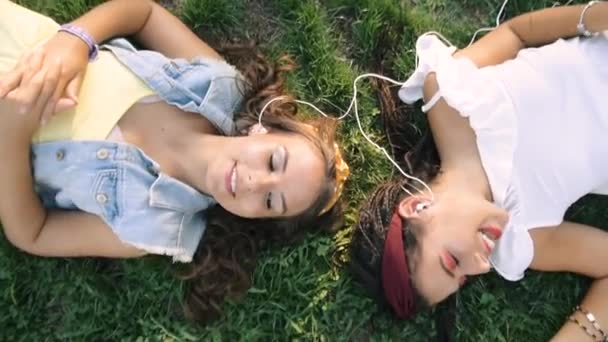 Two girlfriends / sisters are lying on the grass, listening to music on headphones. Two brunettes lie on the grass in the park, relax, sing. - Кадри, відео