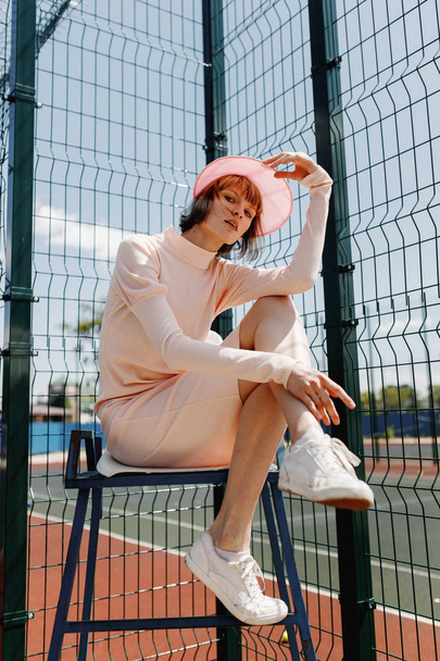 Stylish girl in a gently pink dress and pink visor sitting on a chair behind fence of the lattice at the sports field on a summer sunny day - Foto, afbeelding