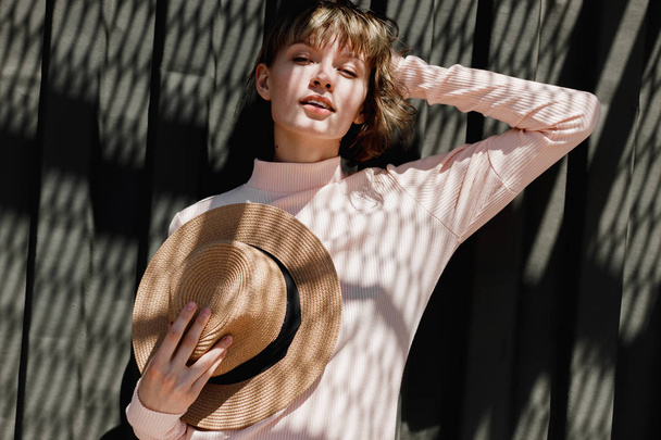 Stylish girl in a gently pink dress and straw hat standing with the straw hat in her hand next to a dark metal fence in the sunlight with reflected shadows - Foto, afbeelding