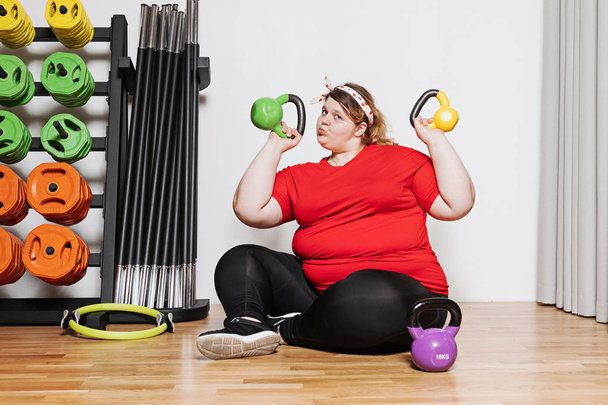 Fat and funny girl dressed in the sports wear is working out with dumbbells in the gym next to other sports equipment - Foto, Bild