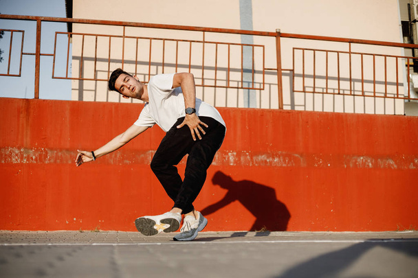 Stylish young guy dressed in jeans and white t-shirt is dancing brakedance in the street against a painted concrete wall in the sunny day - 写真・画像