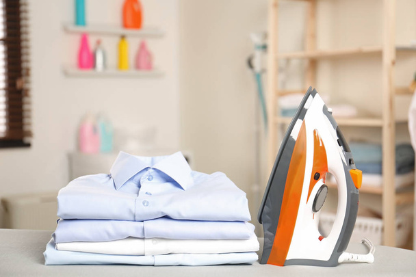 Modern electric iron and folded clothes on board in laundry room - Photo, image