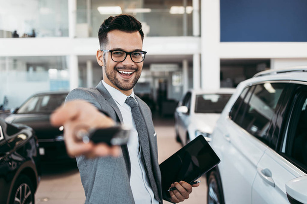 Good looking, smiling salesman poses in a car salon or showroom. He stands with outstretched hand and holds the keys. - Foto, afbeelding