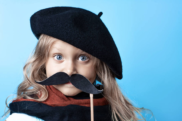 Funny girl in black beret, scarf and mustache on a stick on blue background - Zdjęcie, obraz
