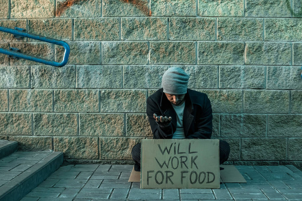 A man, homeless, a man asks for alms on the street with a sign will work for food. Concept of homeless person, addict, poverty, despair. - Photo, image